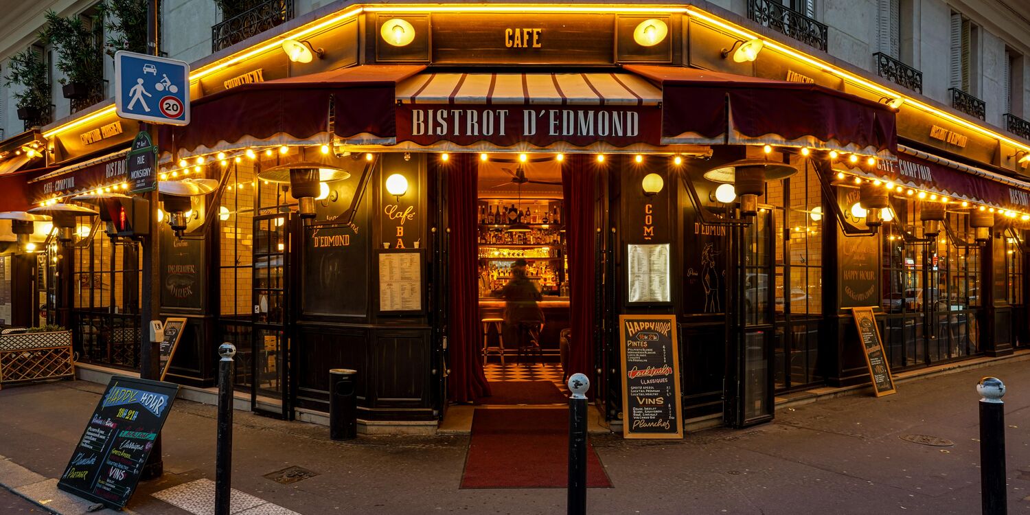Bistrot d’Edmond at night — corner of rue Monsigny, illuminated awnings and string lights, the rue Monsigny street sign on the left