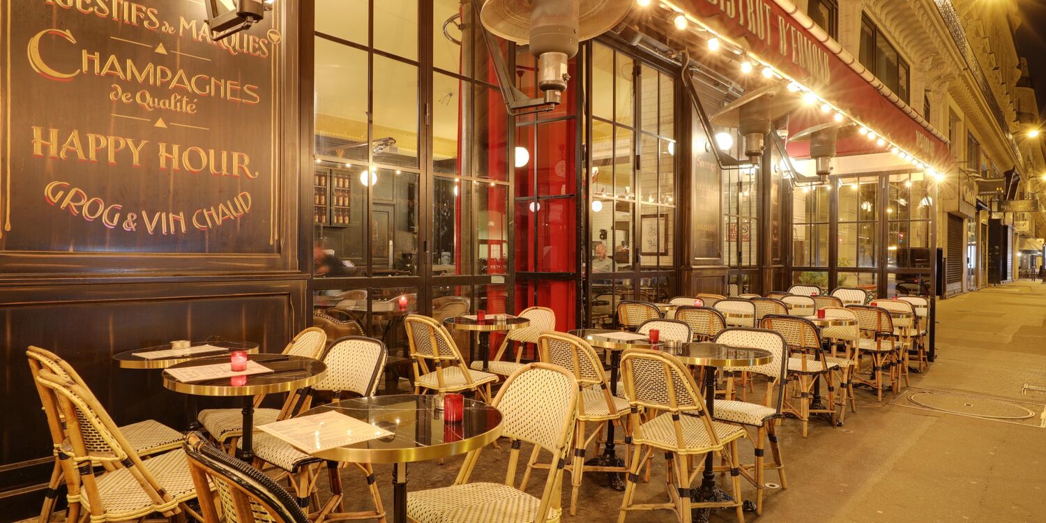 Bistrot d’Edmond terrace at night — Parisian rattan chairs, marble café tables, candles, gold lettering Champagnes Happy Hour Grog & Vin Chaud