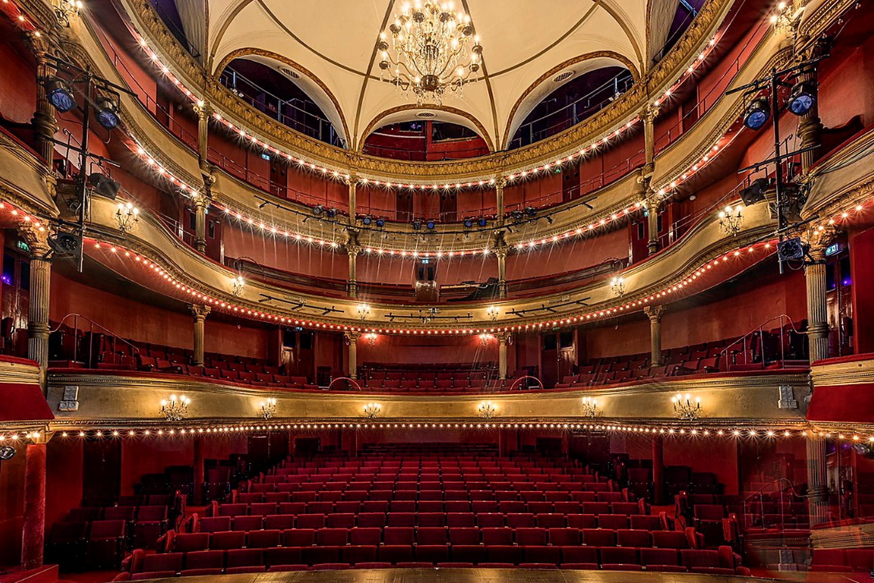 Théâtre des Bouffes-Parisiens interior — Italian-style auditorium seen from the stage, three tiers of gilded balconies trimmed with warm bulb lights, crystal chandelier under the painted dome and rows of crimson velvet seats, Paris 2e